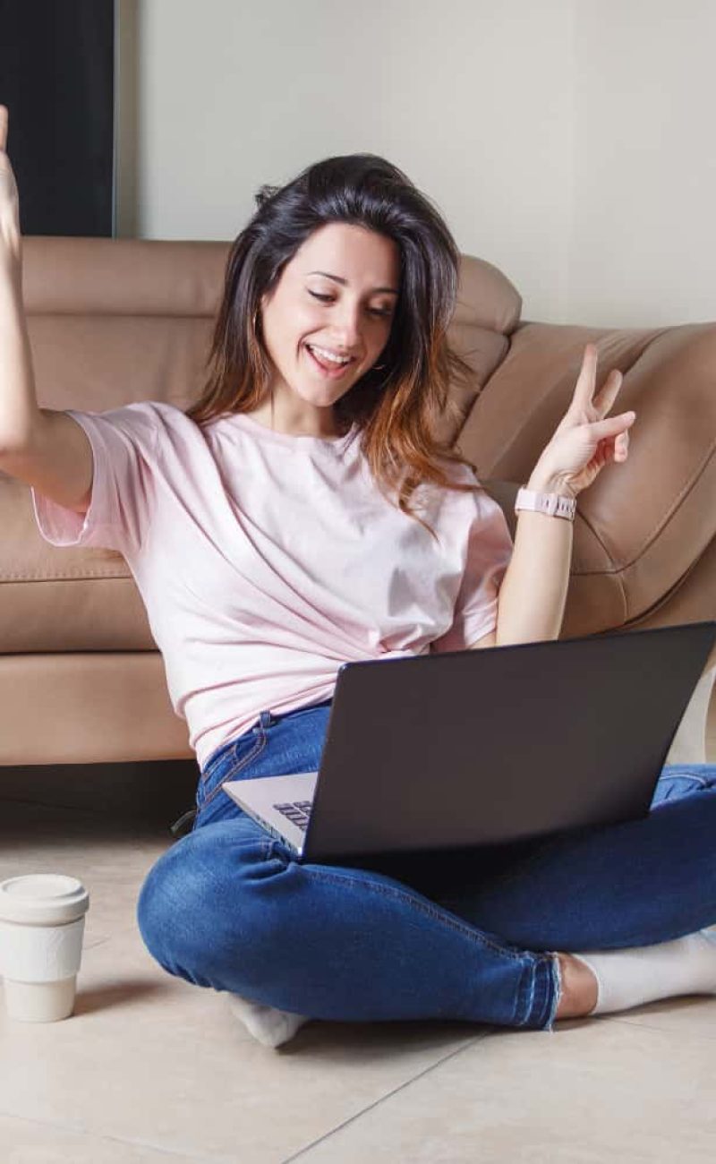young-women-seats-floor-home-with-laptop-show-victoria-gesture-with-fingers-smile-min